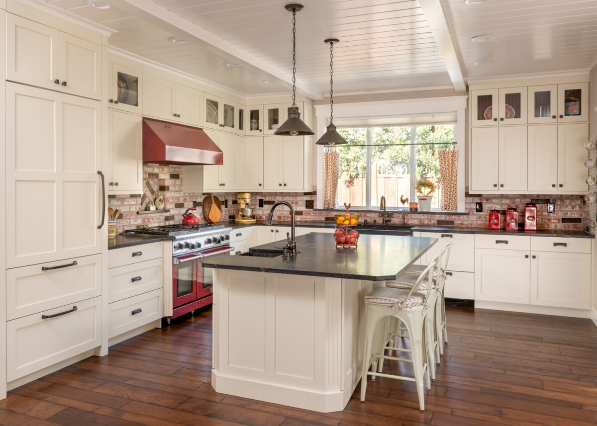 A modern farmhouse kitchen design with white painted cabinets, red brick backsplash, red accents, wood floors and contrasting dark black granite countertops.