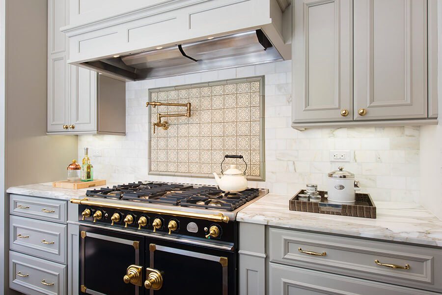 This gray, black, and gold kitchen design has a beautiful cook zone area with functional storage around the range. A large wood hood offers light and ventilation. The cabinets around the range include storage accessories for pots & pans, baking sheets, and cooking utensils.