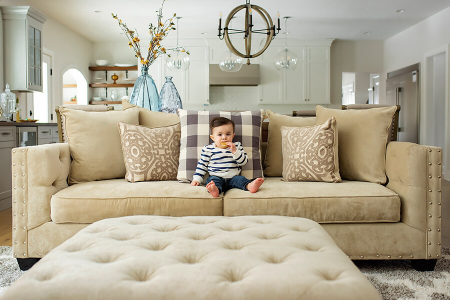 An adorable little toddler is sitting on a living room sofa in front of the new kitchen remodel with white painted kitchen cabinets from Dura Supreme Cabinetry. Undergoing a kitchen or bathroom remodel is a complex process, and there are many things for you to think about as a homeowner in order to ensure the smoothness and success of the project. Among these many considerations is how you’re going to live comfortably during the remodel itself.