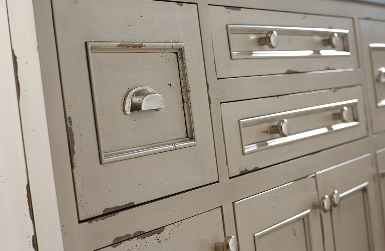 A distressed cabinet finish. This finish technique is hand-detailed by a finish artisan to create a weathered, aged, or antiqued appearance. Here shows a set of painted inset cabinets with a distressed, chipped paint look. This finish is from the Heritage Paint Collection of finishes by Dura Supreme Cabinetry.