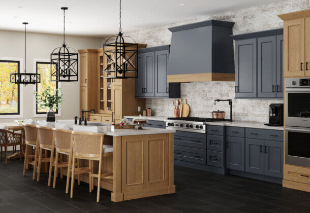 A remodeled kitchen with a modern take on the Old English interior design style. This design features deep blackish navy blue painted cabinets around the cooking space and on the wood hood surrounded by light stained cherry cabinets and wood accents. The kitchen cabinets use a framed construction method with full overlay door that have a flat panel style.