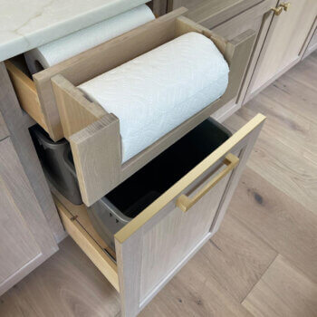 A pull-out trash bin cabinet with a paper towel roll dispenser in the drawer above.