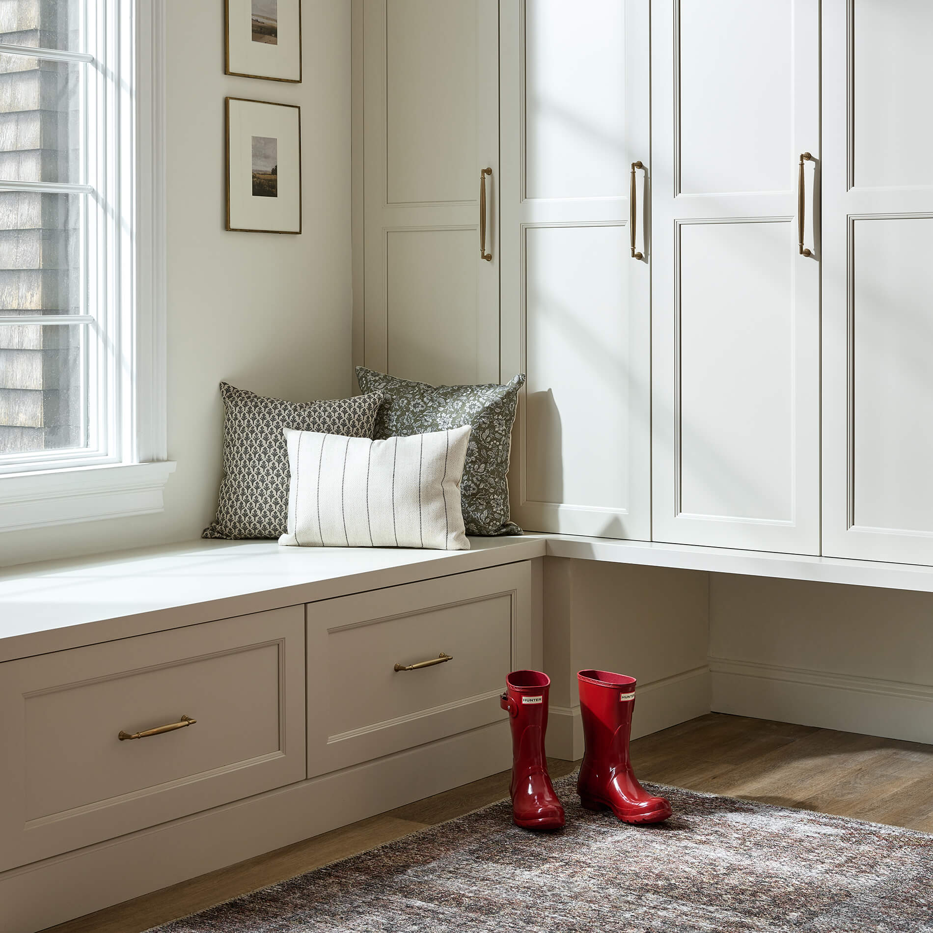 A corner entryway space with mudroom lockers and a built-in boot bench in front of a window featuring a trending classy cabinet door style that has more detail than a typical shaker door. The Meridien door style by Dura Supreme.